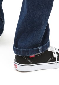 Baskets noires en toile avec lacets blancs et semelle en caoutchouc, assorties à un jean en denim bleu foncé avec revers. Logo Vans visible sur la chaussure et le jean.