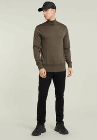 Maglione a collo alto marrone con orlo a coste, abbinato a pantaloni neri slim-fit e scarpe sportive nere, completato con un cappellino nero a fantasia.