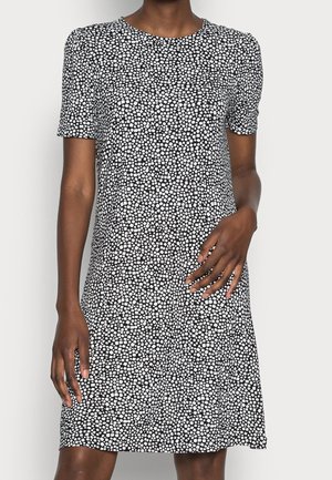 Frau trägt knielanges schwarzes Kleid mit kurzen Ärmeln und weißem, unregelmäßigem Punktmuster, steht vor hellem, einfarbigem Hintergrund.