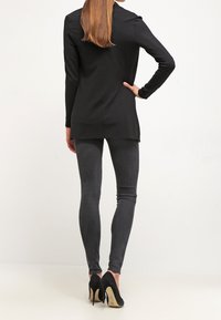 Svart långärmad topp med figurnära design och något förlängd bak. Bärs med mörkgrå leggings och svarta högklackade skor med gulddetaljer.