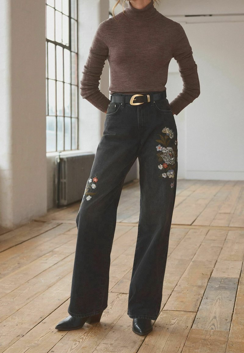 Femme portant un col roulé marron rentré dans un jean noir large avec broderies florales, ceinture noire et bottes pointues noires.