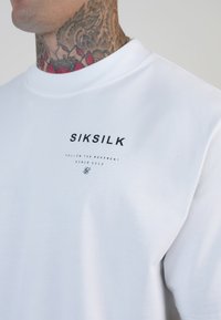 Valge puuvillane T-särk lühikeste varrukatega, millel on must logo teksti "SIKSILK" ja väiksem tekst allpool. Sile tekstuur, ümmargune kaelus.