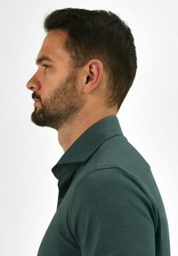 Donkergroene polo met kraag, gemaakt van een gladde stof, draag door een man in profiel. Heeft een getailleerde snit en korte mouwen.