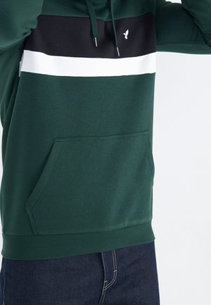 Mand iført mørkegrøn hættetrøje med sorte og hvide vandrette striber og kængurulomme, kombineret med mørkeblå jeans.