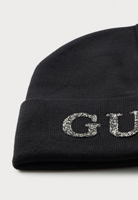 Svart strikket lue med brett som har store sølvfargede rhinestone-bokstaver som staver "GU".