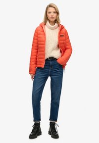 Giacca puffer arancione con cappuccio, chiusura con zip e polsini a coste, abbinata a un maglione di lana crema e jeans blu. Stivali neri con lacci completano il look.