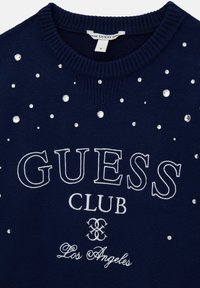Pull en maille bleu marine à col rond, orné de strass argentés et de texte brodé en blanc indiquant "GUESS CLUB Los Angeles".