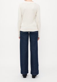 Femme portant un pull crème à manches longues et un jean large bleu foncé, debout de dos sur un fond blanc.