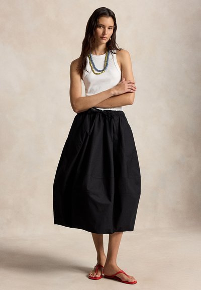 Γυναίκα που φορά λευκό αμάνικο τοπ, στρωματοποιημένα κολιέ με χάντρες, μαύρη μίντι φούστα και κόκκινα σανδάλια με χιαστί λουριά, στέκεται με τα χέρια σταυρωμένα.