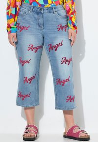 Jeans cortos de color azul claro con texto rojo bordado de "Ángel" disperso por toda la prenda. Combinados con sandalias rosas con bordes festoneados.