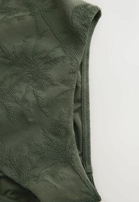 Tissu vert olive avec des motifs de feuilles de palmier texturés, montrant un gros plan du bord sans manches d'un vêtement et des détails de couture.