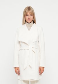 White wrap coat in soft fabric with a shawl collar, long sleeves, and a tied belt; features side pockets and a clean, minimalist design.