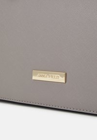Gros plan d’un sac gris texturé avec une étiquette métallique rectangulaire dorée gravée « ANNA FIELD » cousue au bord inférieur.