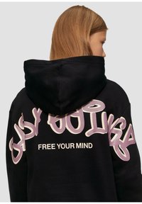 Hoodie noir avec une capuche, portant un grand texte en graffiti violet "EASY GOING" et "FREE YOUR MIND" au dos. Matière douce avec une coupe décontractée.
