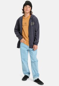 Navy zip-up hoodie over a mustard T-shirt with logo, light blue jeans, and black slip-on shoes. Model poses with one hand in pocket.