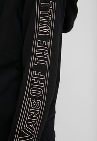 Sweat-shirt noir à manches longues, arborant un logo "VANS OFF THE WALL" en rose pâle sur le bras droit. Texture lisse.