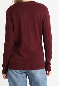 Pull en maille bordeaux avec un col rond, des manches longues et un ourlet côtelé. Le tissu semble doux et cintré à la taille.