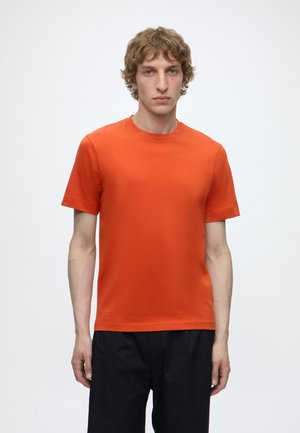 Ung mand med krøllet lys brun hår iført ensfarvet lys orange kortærmet t-shirt og sorte bukser, står mod hvid baggrund.