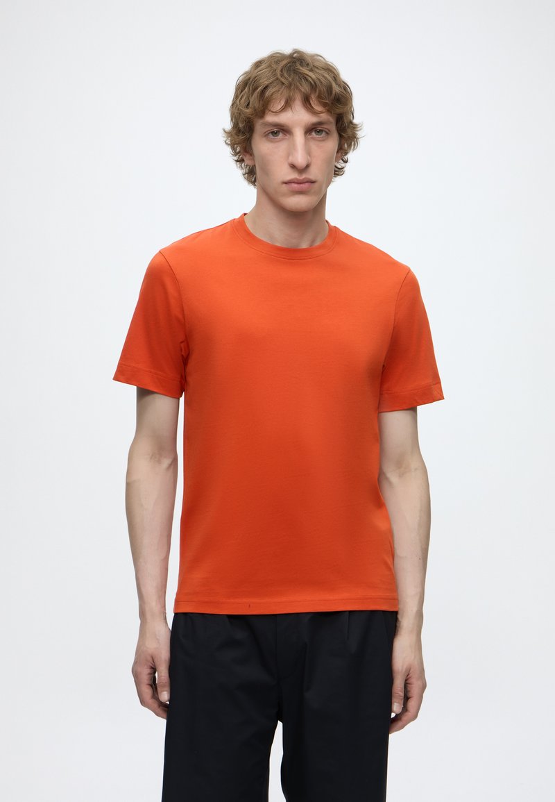 Jeune homme aux cheveux châtain clair bouclés, portant un t-shirt uni orange vif à manches courtes et un pantalon noir, debout devant un fond blanc.