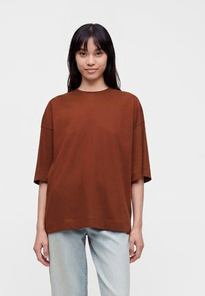 Jeune femme aux cheveux raides et sombres portant un haut marron ample à manches courtes et un jean bleu clair, debout devant un fond uni.