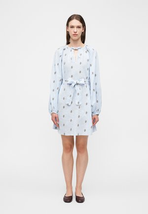 Donna in piedi che indossa un vestito a righe azzurre con stampe di api, maniche lunghe, collo annodato, fiocco in vita e scarpe basse marroni.