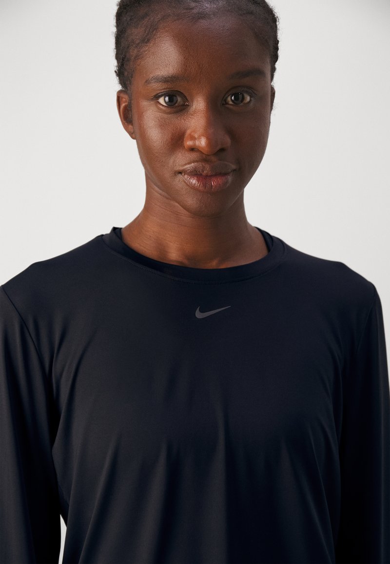 Camiseta deportiva de manga larga negra hecha de tejido elástico, con un cuello redondo y un logo reflectante de Nike en el pecho. Textura suave.