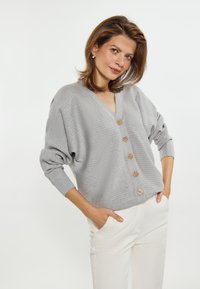 Cardigan din tricot ribbed de culoare gri deschis, cu decolteu în V și cinci nasturi din lemn pe față. Are mâneci supradimensionate și croială lejeră.