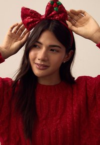 Rotes Haarband mit Pailletten, großer Schleife und grünen Stechpalmenakzenten. Modell trägt einen strukturierten roten Pullover mit Zopfmuster. Hintergrund mit Weichzeichnung.