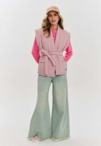 Różowa, pikowana kamizelka z paskiem, z dwiema przednimi kieszeniami. Noszona na jasnoróżowym swetrze i sparowana z szerokimi, jasnoniebieskimi spodniami.