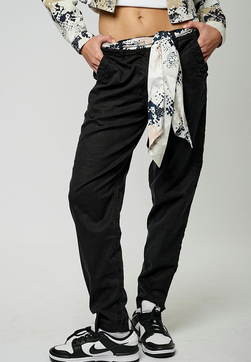 Pantalones negros de corte suelto con cintura elástica, que presentan un cinturón de lazo blanco con patrón y tobillos acampanados, combinados con zapatillas negras.