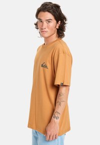 Brown short-sleeve t-shirt with a rounded neck, featuring a black wave logo on the chest and a small label on the side seam. Casual fit.