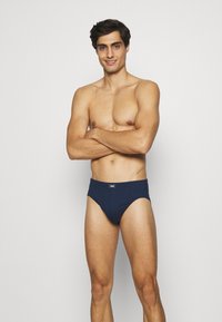 Námorně modré trenky vyrobené z měkké bavlny, s hladkým povrchem a elastickým pasem. Design zahrnuje jednoduchou štítkovou logo nášivku na přední straně.