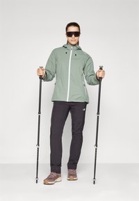 Geacă impermeabilă verde deschis cu glugă, pantaloni de drumeție negri, încălțăminte de drumeție maro, ținând bețe de trekking și purtând ochelari de soare supradimensionați.