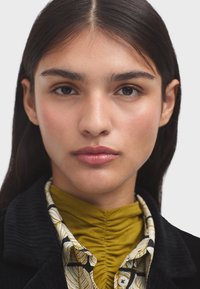 Portrait en gros plan d'une personne avec des cheveux foncés, portant une veste en velours côtelé noir sur une chemise verte à fronces et à motifs avec des accents blancs.