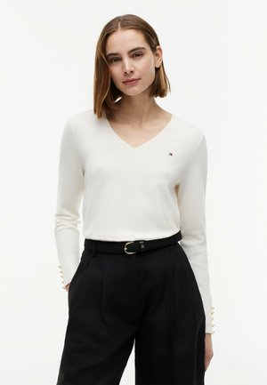 Vrouw met steil bruin haar draagt een witte V-hals trui met klein logo en zwarte hoge taille broek met riem, handen in de zakken.