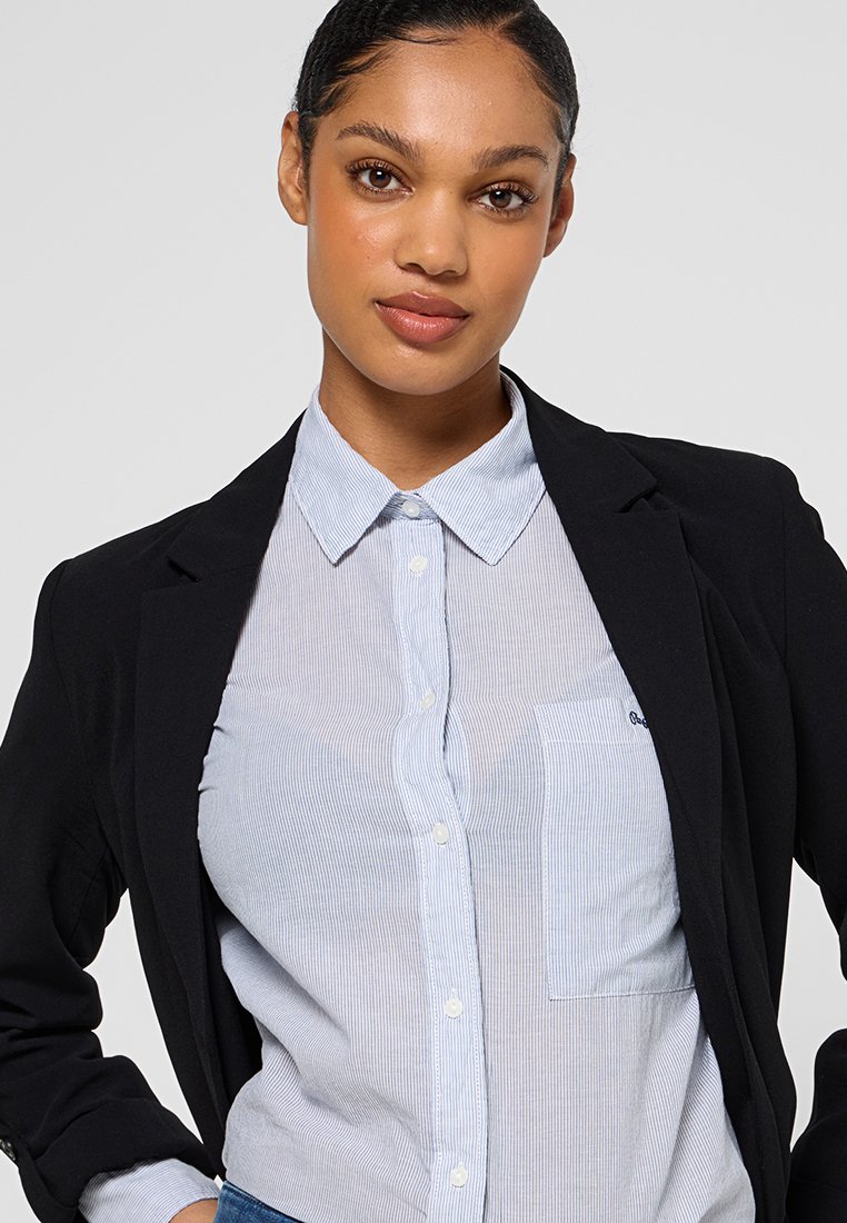 Blazer noir sur une chemise rayée bleu clair avec une poche. La chemise présente un col classique et une fermeture à boutons. Texture lisse.