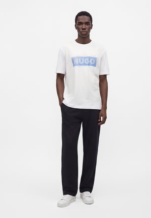 Νέος άνδρας στέκεται φορώντας λευκό T-shirt με μπλε λογότυπο HUGO, μαύρο παντελόνι και λευκά αθλητικά παπούτσια, σε απλό λευκό φόντο.