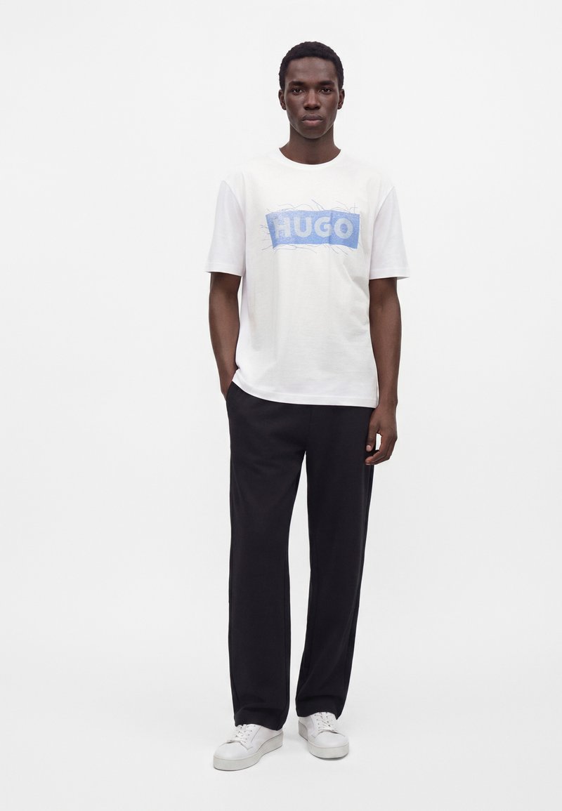 Νέος άνδρας στέκεται φορώντας λευκό T-shirt με μπλε λογότυπο HUGO, μαύρο παντελόνι και λευκά αθλητικά παπούτσια, σε απλό λευκό φόντο.
