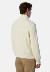 Maglione bianco a costine con colletto alto e maniche lunghe. Design a maglia testurizzato che si estende fino all'orlo. Indossato sopra pantaloni marrone chiaro.