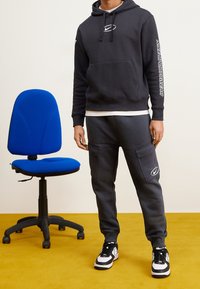 Svart hoodie med logotyp, grå sweatpants med fickor och svarta och vita sneakers. Blå ergonomisk stol på gul golv.