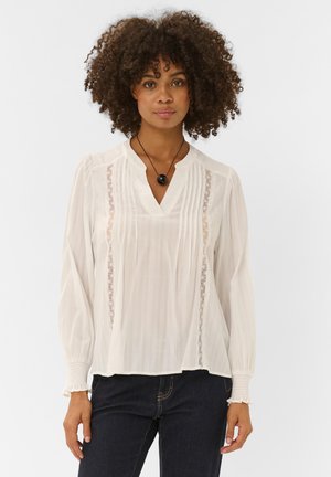 Femme aux cheveux bouclés portant un chemisier blanc à manches longues avec des détails en dentelle et un jean foncé, debout devant un fond uni.