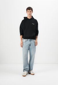 Hoodie noir avec une poche avant et un logo, associé à un jean clair ample et des baskets blanches avec des accents bruns.