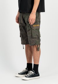 Zaļi kamuflāžas šorti, izgatavoti no izturīga auduma. Iespējas: sānu kabatas, gumijas apakšmalas un dzeltena etiķete uz kabatas.