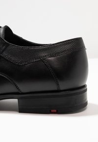 Schwarzer Lederschuh mit texturiertem schwarzen Obermaterial, glatter Oberfläche, niedrigem Absatz und einem kontrastierenden roten Akzent an der Sohle.