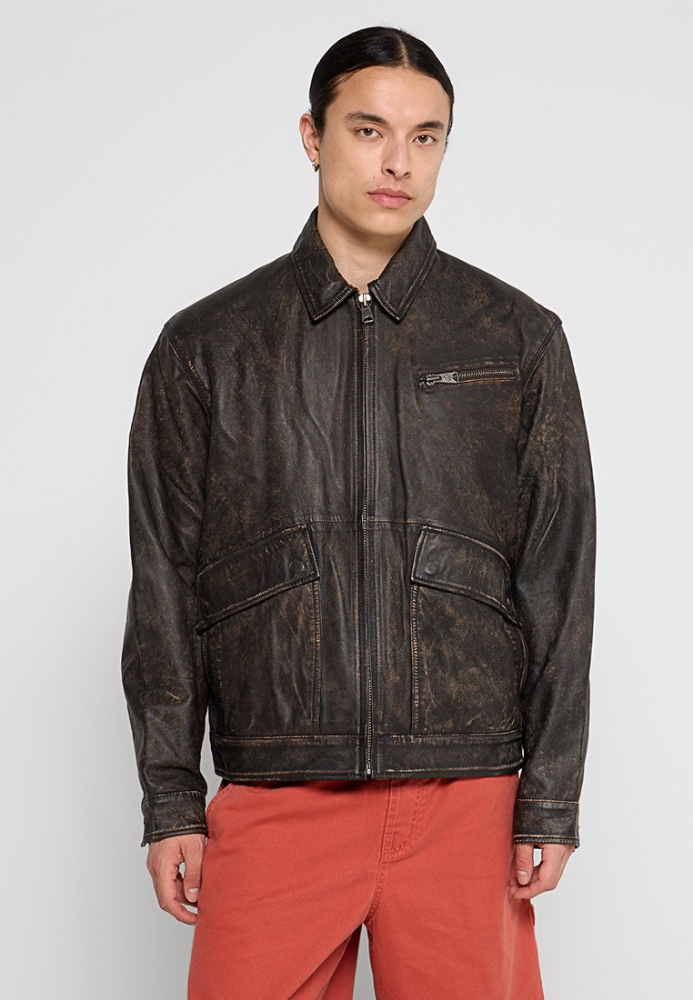 Brązowa skórzana kurtka z zamkiem z przodu, dwiema dużymi kieszeniami z przodu i małą kieszonką na piersi. Tekstura jest lekko przetarta.