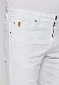 Jeans de mezclilla blanca con cremallera, bolsillos frontales y una pequeña etiqueta de marca marrón. Presenta herrajes en tono plateado y una textura suave.