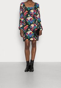 Vrouw in een zwart bloemenprint lange mouwen jurk, met een zwarte clutch, gecombineerd met zwarte enkellaarsjes, staande tegen een effen achtergrond.