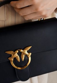 Čierna kožená kabelka s zlatým kruhovým zapínaním a dvoma precízne vypracovanými akcentmi vtákov, hladkou textúrou a úhľadným šitím.