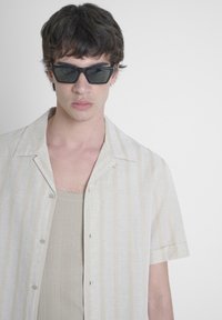Camisa de manga corta a rayas en beige claro con líneas verticales sutiles, combinada con unas gafas de sol rectangulares oscuras y una camiseta sin mangas beige.