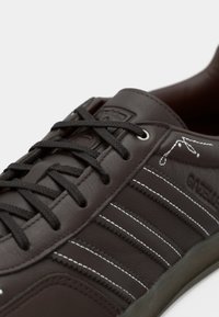 Bruine leren sneaker met donkere veters, witte strepen en merkdetails. Het ontwerp heeft een glanzende afwerking en een rond teenmodel.